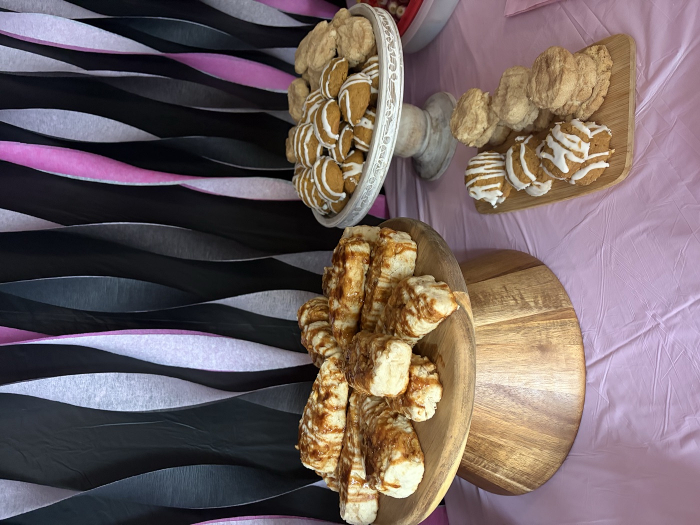 Sticky buns on a wooden cake stand next to iced cookies on a pedestal and scones on a board