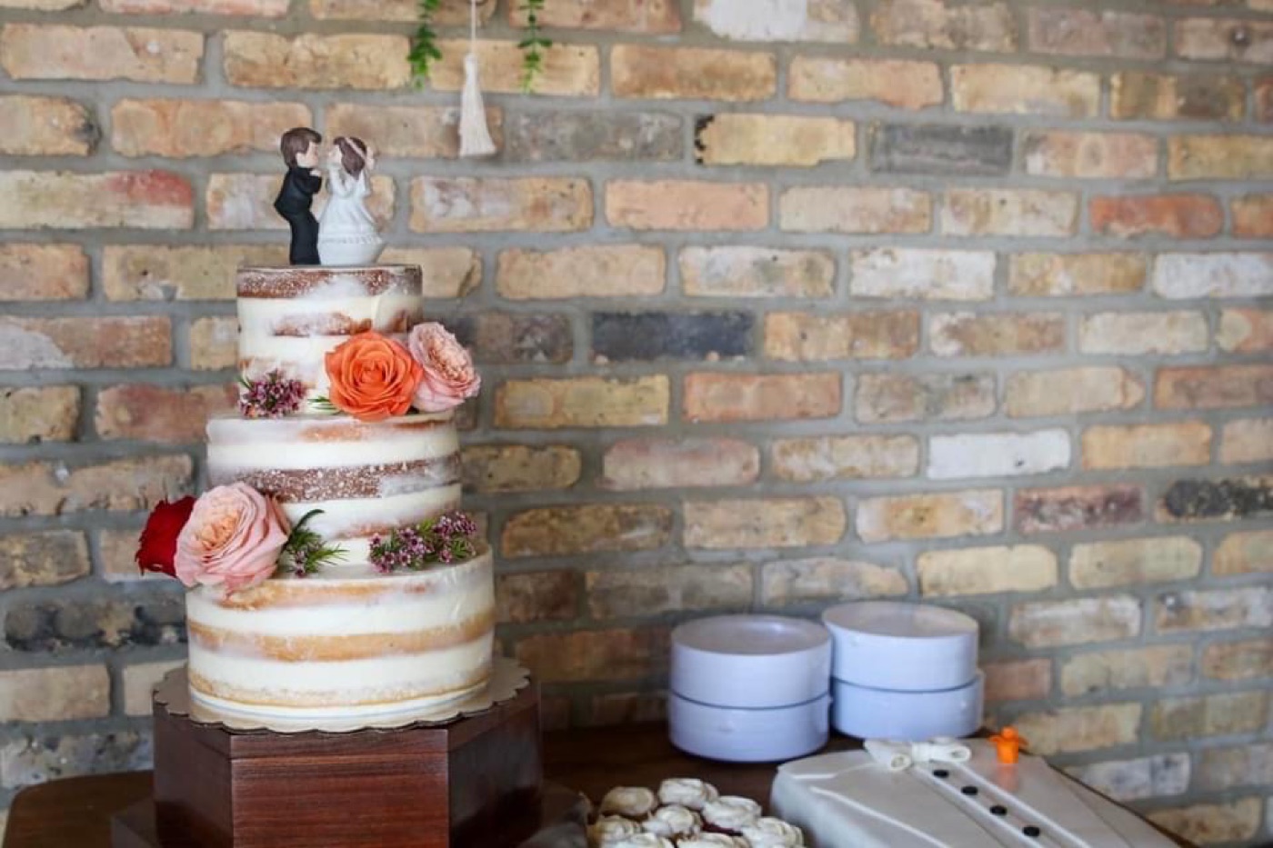 Three-tier naked wedding cake with roses against a brick wall, beside a groom&rsquo;s cake and cookies